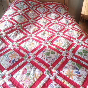 May include: A floral quilt with a red diamond pattern. The quilt features a white background with colorful flowers and green leaves. The red borders create a striking contrast, adding visual interest to the bedding.