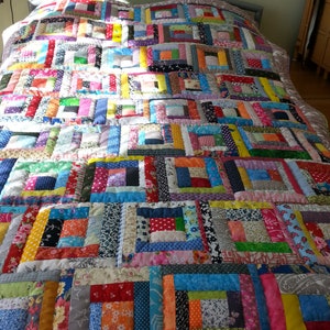 Könnte beinhalten: Eine bunte Patchwork-Decke mit einem Blockhausmuster. Die Decke besteht aus verschiedenen Stoffen in den Farben Rosa, Blau, Grün, Gelb und Rot. Die Decke liegt auf einem Bett mit einem weißen Laken darunter.