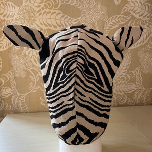 May include: A black and white zebra-print hooded hat with ears. The hat is made of fabric and is sitting on a white mannequin head.