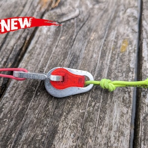 Puede incluir: Primer plano de un clip metálico plateado y rojo con un cordón verde sujeto. El clip está conectado a un cordón rosa. El clip está sobre una superficie de madera.