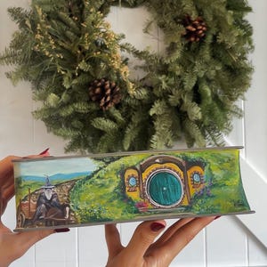 May include: A hand-painted wooden box with a scene from The Lord of the Rings. The box features a hobbit hole with a round green door and a path leading to it. A figure in a blue hat and brown cloak is walking on the path.