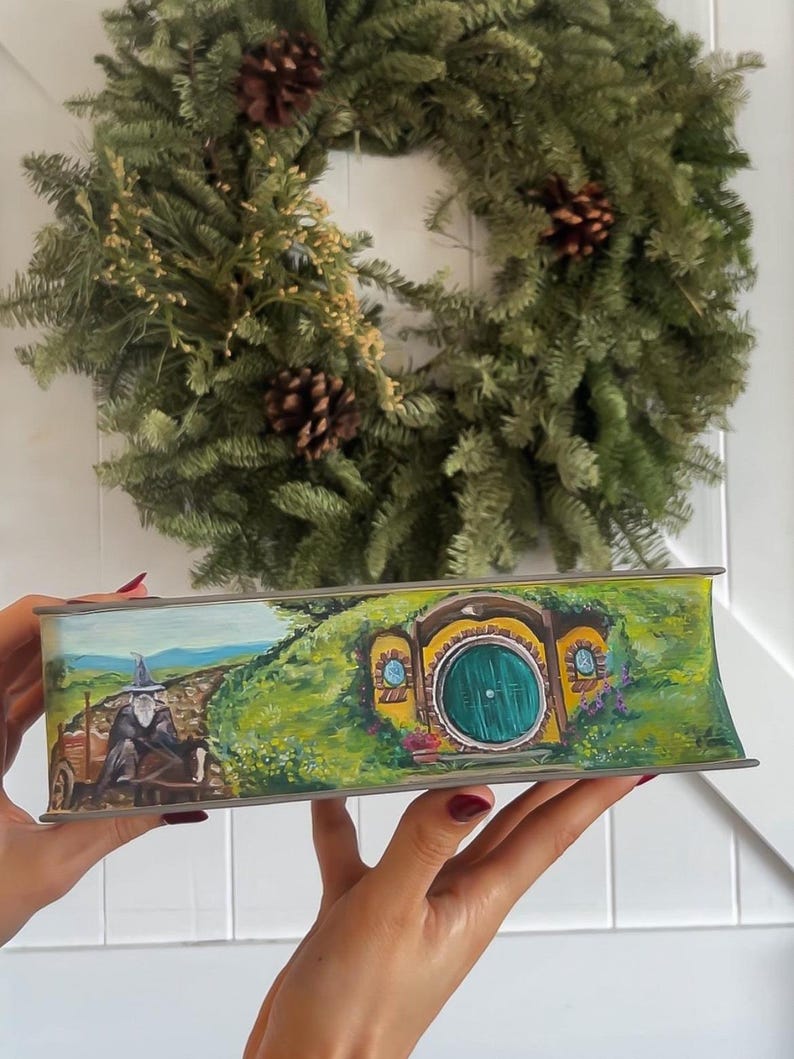 May include: A hand-painted wooden box with a scene from The Lord of the Rings. The box features a hobbit hole with a round green door and a path leading to it. A figure in a blue hat and brown cloak is walking on the path.