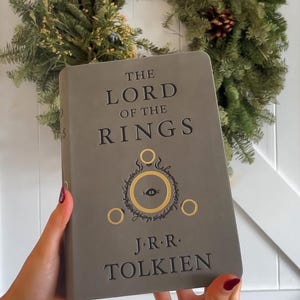 May include: A gray hardcover book with the title "The Lord of the Rings" and the author "J.R.R. Tolkien" printed on the front cover. The book features a gold embossed illustration of the One Ring and the Eye of Sauron.