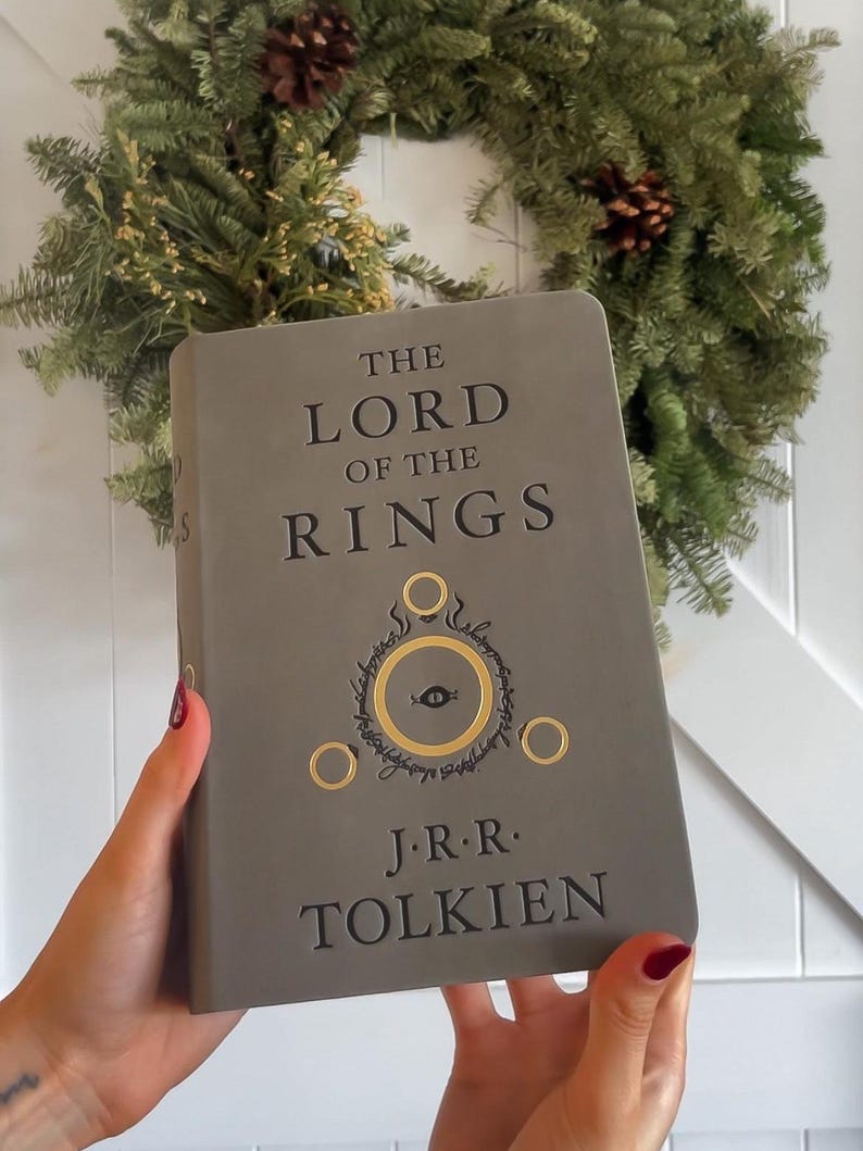 May include: A gray hardcover book with the title "The Lord of the Rings" and the author "J.R.R. Tolkien" printed on the front cover. The book features a gold embossed illustration of the One Ring and the Eye of Sauron.