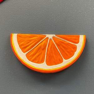 May include: A wooden orange slice with a bright orange interior, white segments, and a yellow and white rind. The slice is set against a gray background.