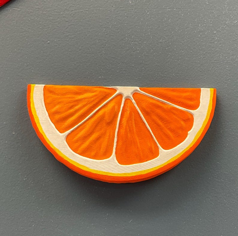 May include: A wooden orange slice with a bright orange interior, white segments, and a yellow and white rind. The slice is set against a gray background.
