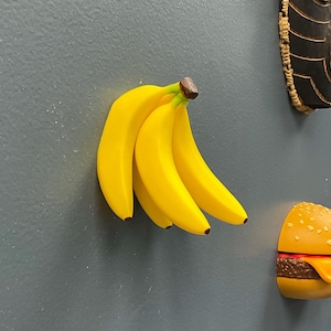 May include: A cluster of four bright yellow banana-shaped magnets with brown stems. The bananas are affixed to a gray surface, with a burger magnet and a carved wooden mask visible in the background. The magnets are likely for the refrigerator.