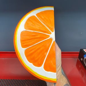 May include: A half-slice of an orange, painted with bright orange segments, white pith, and a yellow and white rind. The artwork is held in a hand, with a dark blue and red background.
