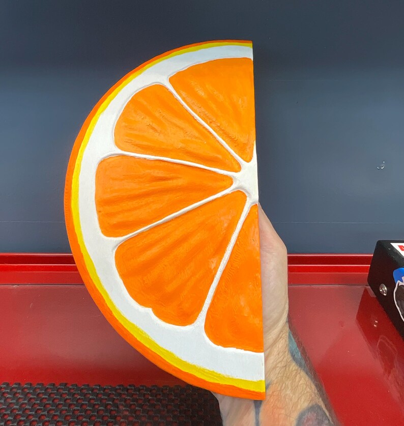May include: A half-slice of an orange, painted with bright orange segments, white pith, and a yellow and white rind. The artwork is held in a hand, with a dark blue and red background.