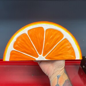May include: A large, half-circle orange slice wall art piece. The artwork features a bright orange interior with white segments and a yellow and white rind. The piece is held by a hand, showcasing the size and detail of the painted design.