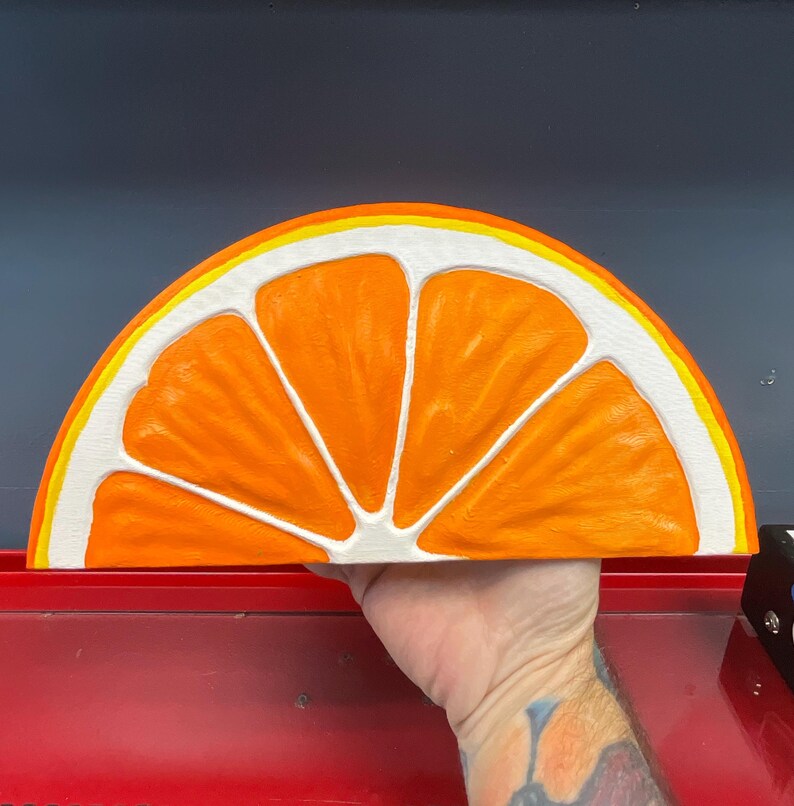 May include: A large, half-circle orange slice wall art piece. The artwork features a bright orange interior with white segments and a yellow and white rind. The piece is held by a hand, showcasing the size and detail of the painted design.
