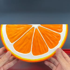 May include: A large, orange, half-slice of a citrus fruit, possibly an orange, with white and yellow accents. The fruit is held by two hands, and the background is a solid, dark blue color.