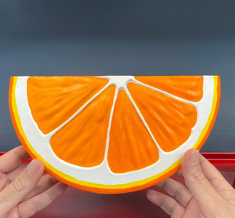 May include: A large, orange, half-slice of a citrus fruit, possibly an orange, with white and yellow accents. The fruit is held by two hands, and the background is a solid, dark blue color.