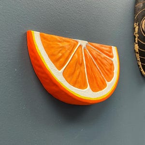 May include: A vibrant, three-dimensional orange slice with a realistic design. The orange features a bright orange exterior, white segments, and a yellow border. The artwork is mounted on a gray surface.