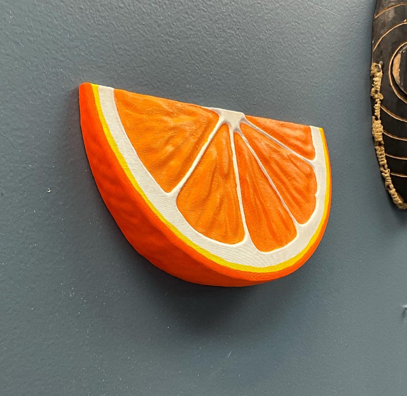 May include: A vibrant, three-dimensional orange slice with a realistic design. The orange features a bright orange exterior, white segments, and a yellow border. The artwork is mounted on a gray surface.