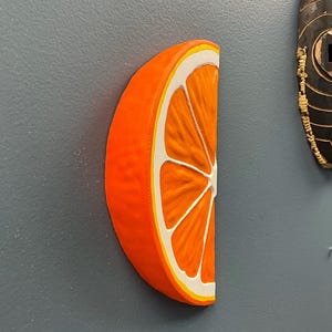 May include: A vibrant orange, half-slice of an orange with white and yellow accents. The fruit is displayed against a gray background. The image showcases a detailed, three-dimensional representation of the citrus fruit.