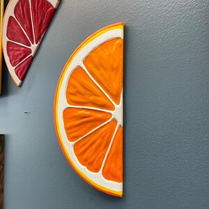 May include: Orange slice wall art with a white and yellow border. The orange slice is painted with a textured, realistic design. The art is mounted on a gray wall. A red grapefruit slice is also visible.