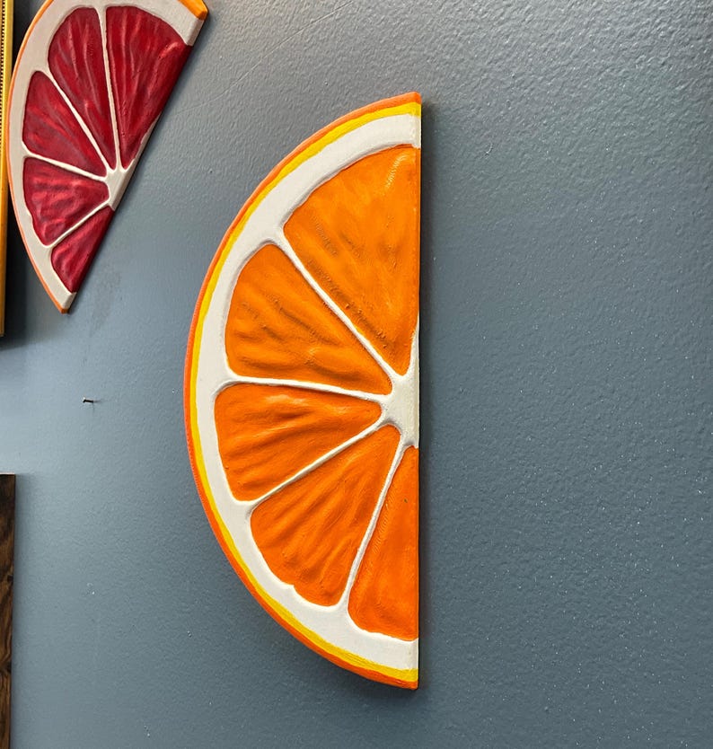 May include: Orange slice wall art with a white and yellow border. The orange slice is painted with a textured, realistic design. The art is mounted on a gray wall. A red grapefruit slice is also visible.