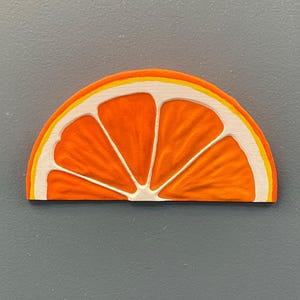 May include: A wooden orange slice with a textured surface. The orange segment is bright orange with white and yellow accents. The slice is a half-circle shape, and the background is a solid gray.