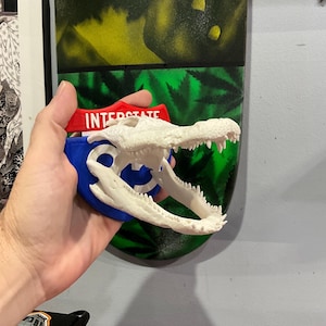 May include: A white 3D printed alligator head with an open mouth, mounted on a blue and white interstate highway sign. The sign has the text "INTERSTATE" in red letters.