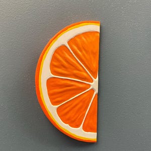 May include: A half-slice of an orange, featuring a bright orange interior with white and yellow accents. The fruit's segments are clearly defined, and the rind is detailed with a yellow and white border. The image is set against a gray background.