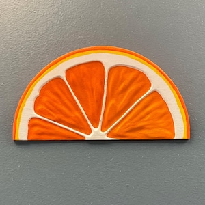 May include: A wooden orange slice with a textured surface. The orange segment is painted in bright orange with white and yellow accents. The artwork is set against a gray background.