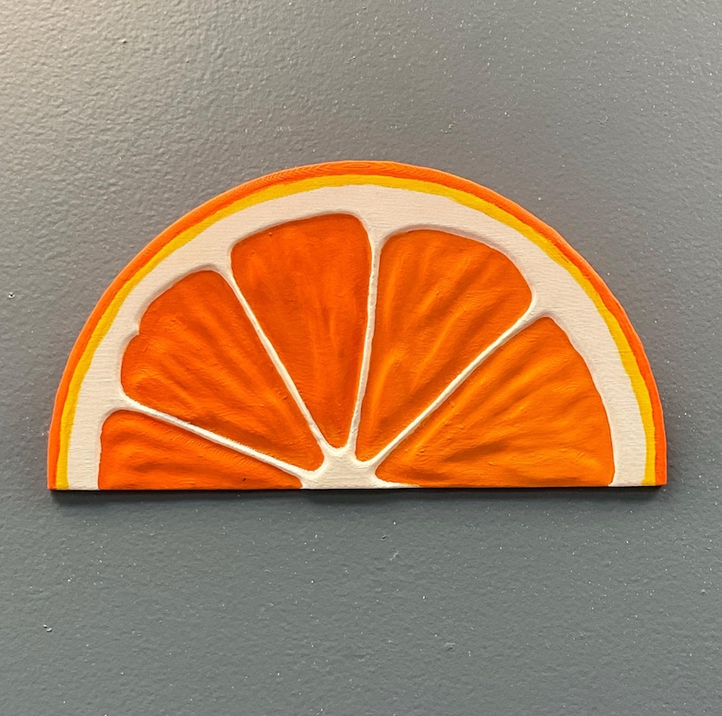 May include: A wooden orange slice with a textured surface. The orange segment is painted in bright orange with white and yellow accents. The artwork is set against a gray background.