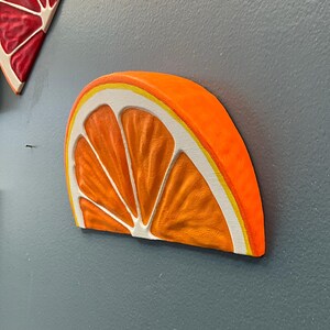 May include: A vibrant orange slice wall art piece with a white and yellow border. The artwork is mounted on a gray wall, showcasing the detailed texture and bright colors of the fruit.