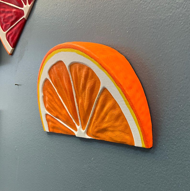 May include: A vibrant orange slice wall art piece with a white and yellow border. The artwork is mounted on a gray wall, showcasing the detailed texture and bright colors of the fruit.