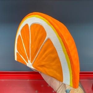May include: A large, three-dimensional orange slice sculpture. The sculpture is painted in bright orange, white, and yellow. The orange slice is a half-circle shape, with the segments clearly defined. The rind is white with a yellow stripe.