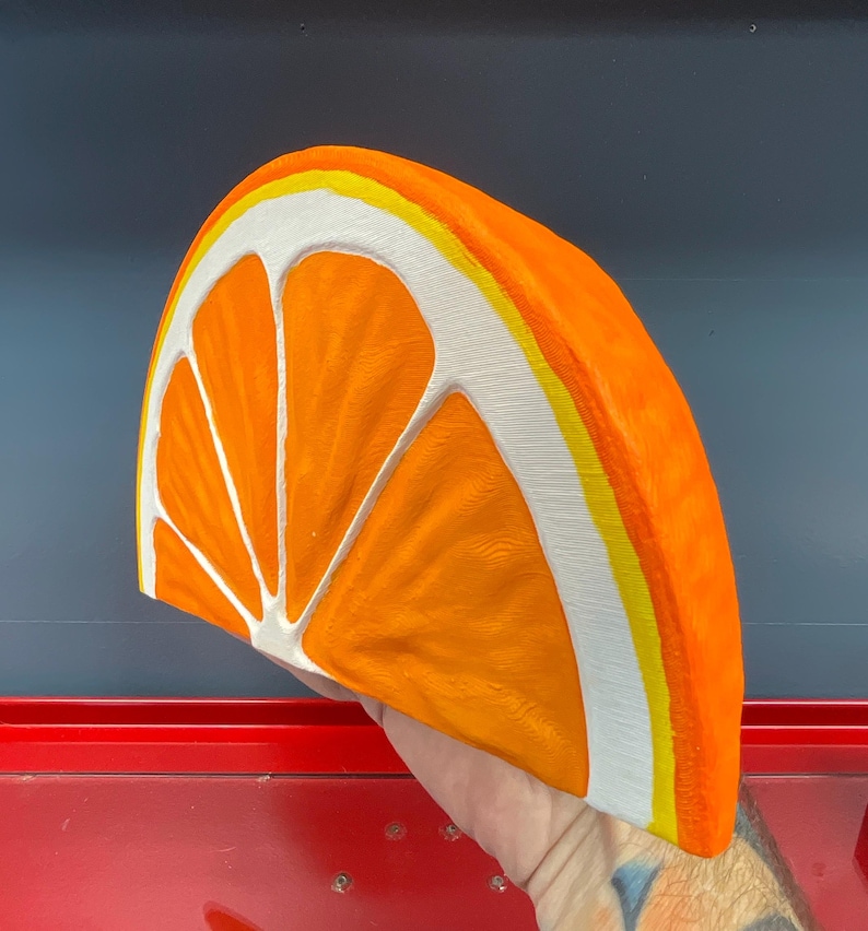 May include: A large, three-dimensional orange slice sculpture. The sculpture is painted in bright orange, white, and yellow. The orange slice is a half-circle shape, with the segments clearly defined. The rind is white with a yellow stripe.