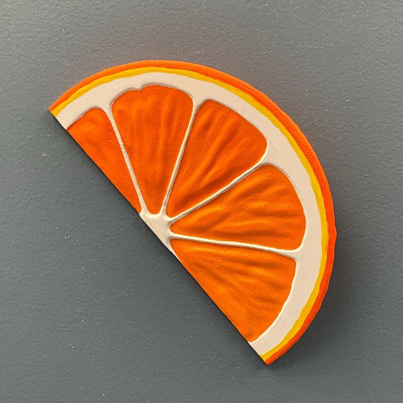 May include: A vibrant orange slice with white and yellow accents. The fruit segment is a half-circle shape, with a textured surface and detailed segments. The artwork is set against a gray background.