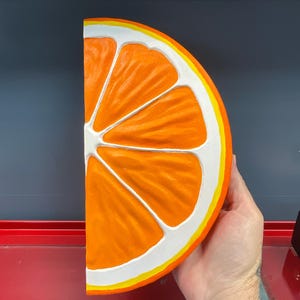 May include: A large, half-cut orange slice with a bright orange interior, white segments, and a yellow and white rind. The fruit is held by a hand, showcasing the vibrant colors and detailed texture of the painted artwork.