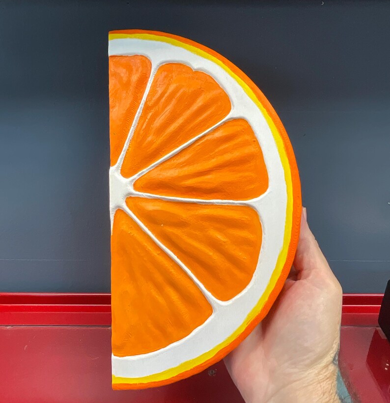 May include: A large, half-cut orange slice with a bright orange interior, white segments, and a yellow and white rind. The fruit is held by a hand, showcasing the vibrant colors and detailed texture of the painted artwork.
