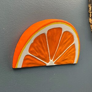 May include: A vibrant, orange, half-slice of an orange, with white and yellow accents. The wooden carving is mounted on a gray wall. The fruit slice is a decorative item.
