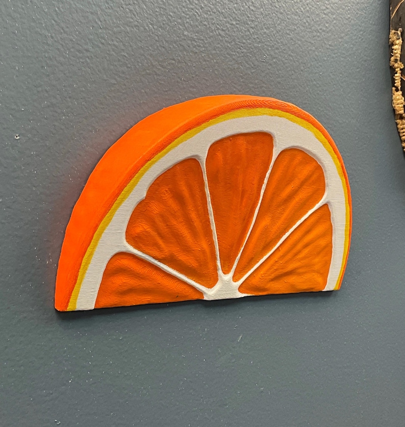 May include: A vibrant, orange, half-slice of an orange, with white and yellow accents. The wooden carving is mounted on a gray wall. The fruit slice is a decorative item.