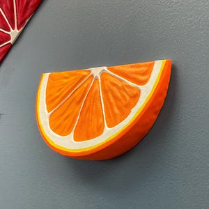 May include: A vibrant orange slice sculpture with white and yellow accents. The artwork is a half-circle shape, with detailed segments and a textured surface. The piece is mounted on a gray wall, suggesting a kitchen or dining room theme.