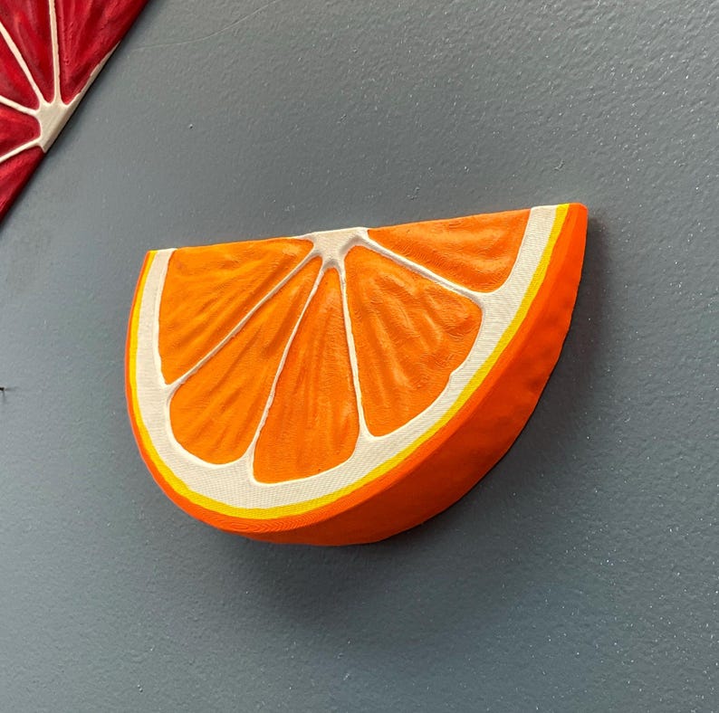 May include: A vibrant orange slice sculpture with white and yellow accents. The artwork is a half-circle shape, with detailed segments and a textured surface. The piece is mounted on a gray wall, suggesting a kitchen or dining room theme.