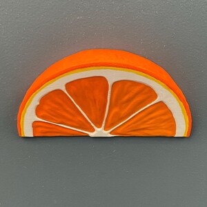May include: A wooden orange slice with a bright orange exterior, a yellow and white rind, and white segments. The artwork is set against a gray background.