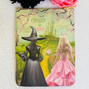 Puede incluir: Una funda para libro con una ilustración caprichosa de dos mujeres, una con un disfraz de bruja negro y la otra con un vestido rosa, caminando por un camino de ladrillos amarillos. El texto "CHANGED for GOOD" está impreso en la funda.