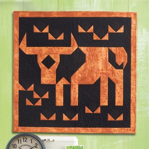 May include: A brown and black quilt with a geometric design of a bull. The quilt is hanging on a green wall with a clock, a white box, and a green plant on a shelf below it.