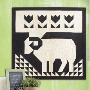 May include: Black and white quilt featuring a stylised ram design. The quilt is framed and hanging on a green wall. A small chalkboard sign with the text "Temperament-voll und furchtlos, wie ein WIDDER" is on a shelf below the quilt.