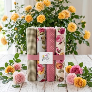 Könnte beinhalten: Ein Bündel aus fünf Stoffrollen mit verschiedenen Mustern, darunter Vichy, Blumen und Punkte, zusammengehalten von einem rosa Band. Das Logo "Quilt Shop" ist zu sehen. Rosen in Rosa und Gelb umgeben das Bündel. Ideal zum Quilten.