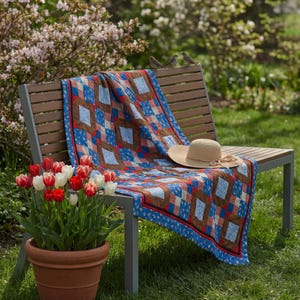 Könnte beinhalten: Ein Patchwork-Quilt mit einem wiederkehrenden Muster aus blauen Blumenquadraten, einfarbigen braunen Rauten und roten Akzenten, drapiert über einer Lattenbank. Ein breitkrempiger Strohhut mit braunem Band liegt darauf, neben einem Tontopf mit Tulpen.