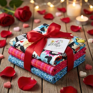 Könnte beinhalten: Ein Stapel aus drei Stoffquadraten, gebunden mit einem roten Band und einer Schleife. Das obere Quadrat hat ein Comic-Design, die Mitte ist rosa kariert und das untere ist blau geblümt. Ein Geschenkanhänger trägt die Aufschrift "Quilt Shop" und "Ruth Baudisch". Rote Rosenblätter und Kerzen umgeben das Geschenk.