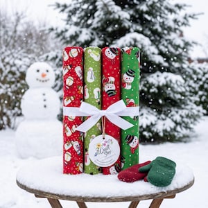 Può includere: Quattro rotoli di tessuto con motivi di pupazzi di neve in rosso e verde, legati insieme da un nastro bianco e un'etichetta circolare con la scritta Quilt Shop Winterwunderland. Un paio di piccole muffole in maglia rossa e verde sul tavolo.