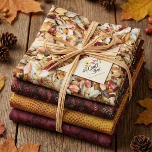 Op de afbeelding: Een stapel herfststoffenbundels vastgebonden met raffia. De bovenste stof heeft een bladerpatroon, met andere stoffen in bordeauxrood en goud. Een label met de tekst "Quilt Shop" is bevestigd. Herfstbladeren en dennenappels omringen de stof.