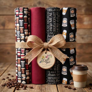 May include: A collection of fabric rolls, tied with twine and a burlap bow. The rolls display coffee cup and food-related patterns. A small coffee cup is on the right. A wooden tag reads "Quilt Shop" and "Ruth Baudisch".