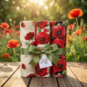 Könnte beinhalten: Drei Stoffrollen mit roten Mohnblumenmustern auf cremefarbenem und schwarzem Hintergrund, gebunden mit einem grünen Band und einer Schleife. Ein Etikett von "Quilt Shop" ist angebracht. Der Hintergrund zeigt Mohnblumen, was auf eine Szene im Freien hindeutet.