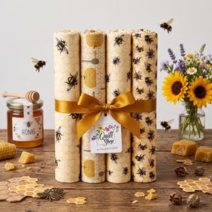 Könnte beinhalten: Vier Stoffrollen mit Bienen- und Wabenmustern, zusammengebunden mit einem goldenen Band und einem Etikett mit der Aufschrift Quilt Shop Ruth Baudisch. Die Stoffrollen sind vertikal angeordnet und zeigen verschiedene Illustrationen von Bienen und Honiglöffeln.